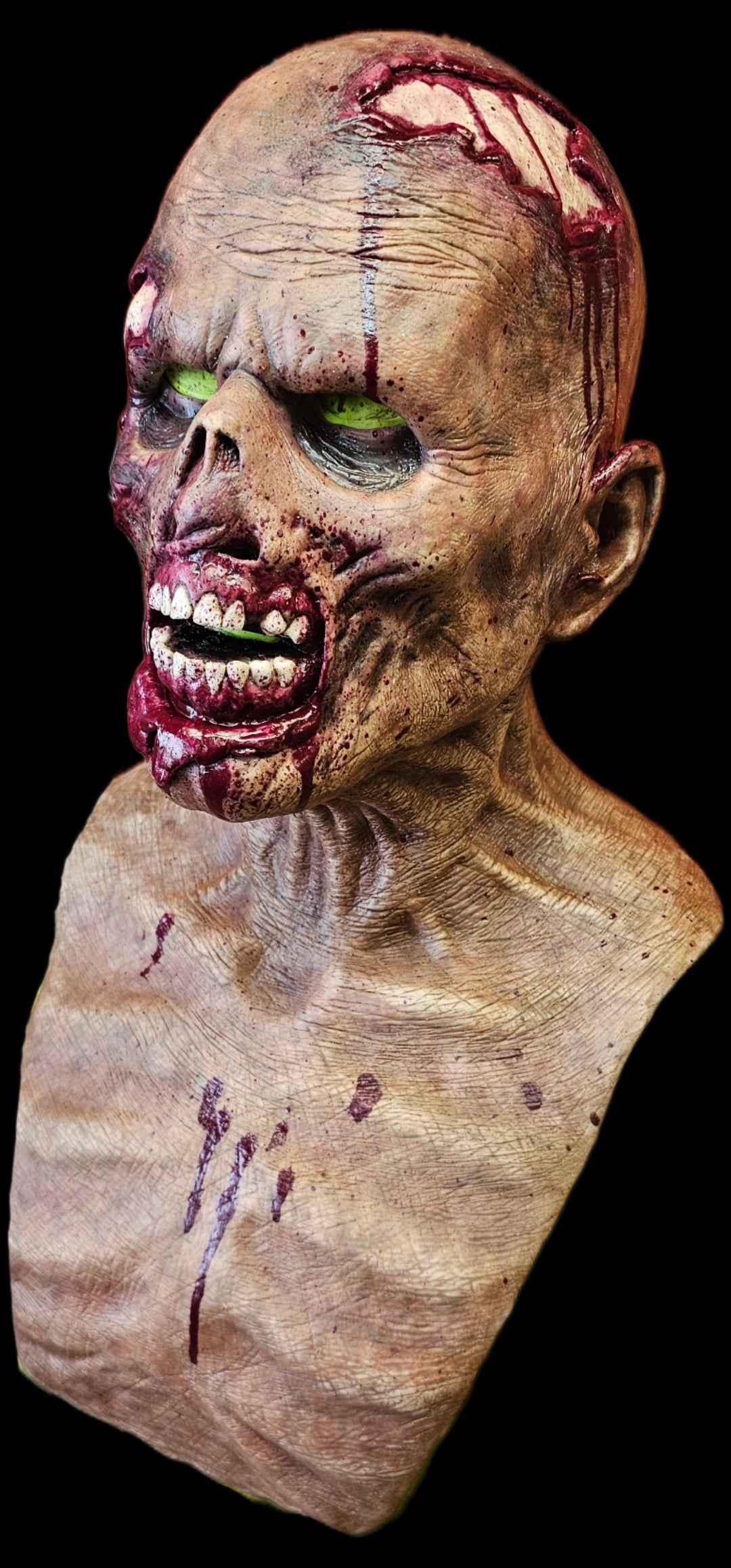 Hyper-realistic silicone zombie mask bust featuring deeply textured decayed skin, glowing green eyes, torn flesh around the mouth with exposed teeth, and blood detailing across the scalp and chest, photographed against a black background.